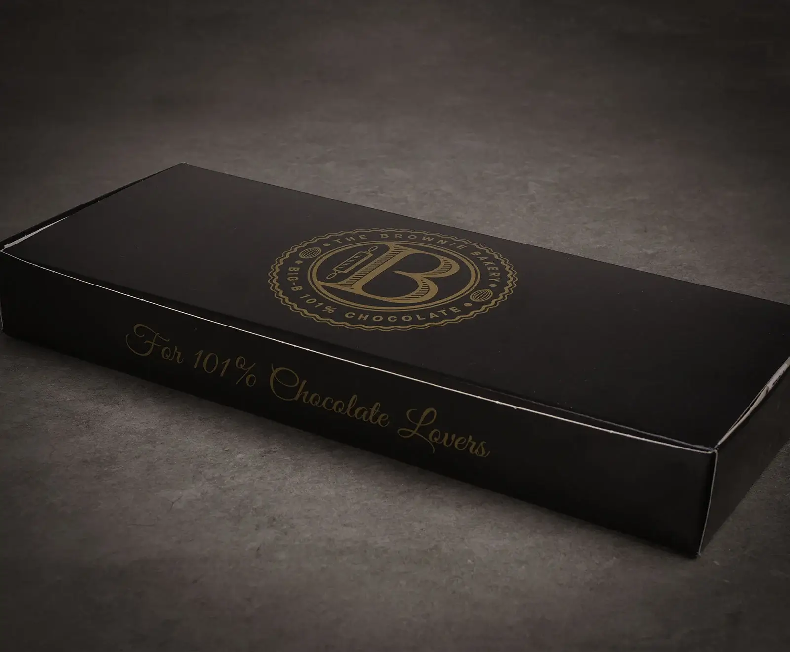 Eine schwarze, rechteckige Schachtel mit goldener Aufschrift. Oben ist ein rundes Logo mit "THE BROWNIE BAKERY" und "BIG B 101% CHOCOLATE" zu sehen. Auf der Vorderseite steht "For 101% Chocolate Lovers".