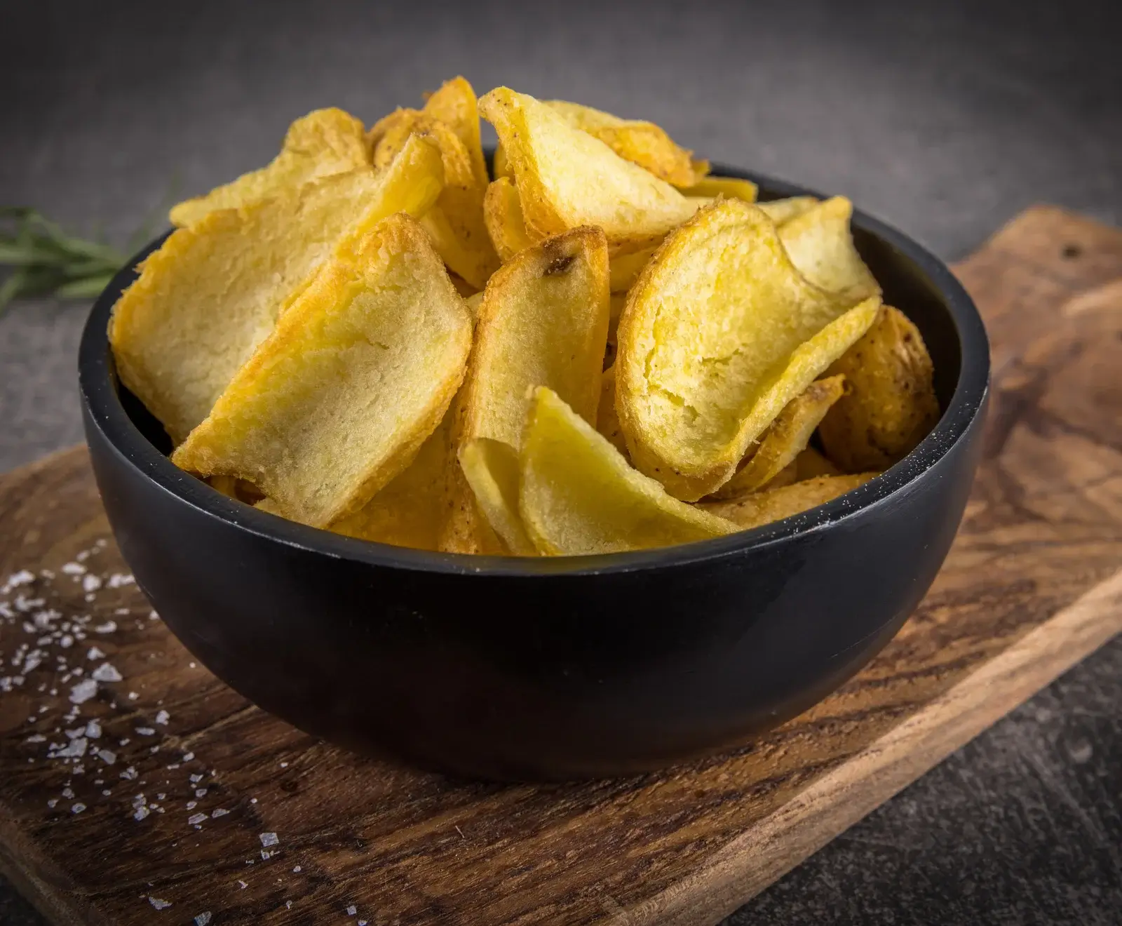 Eine schwarze Schale gefüllt mit goldgelben, rustikalen Kartoffelchips steht auf einem Holzbrett. Grobe Salzkristalle liegen neben der Schale. Im Hintergrund sind dunkle Oberflächen und Rosmarin zu sehen.