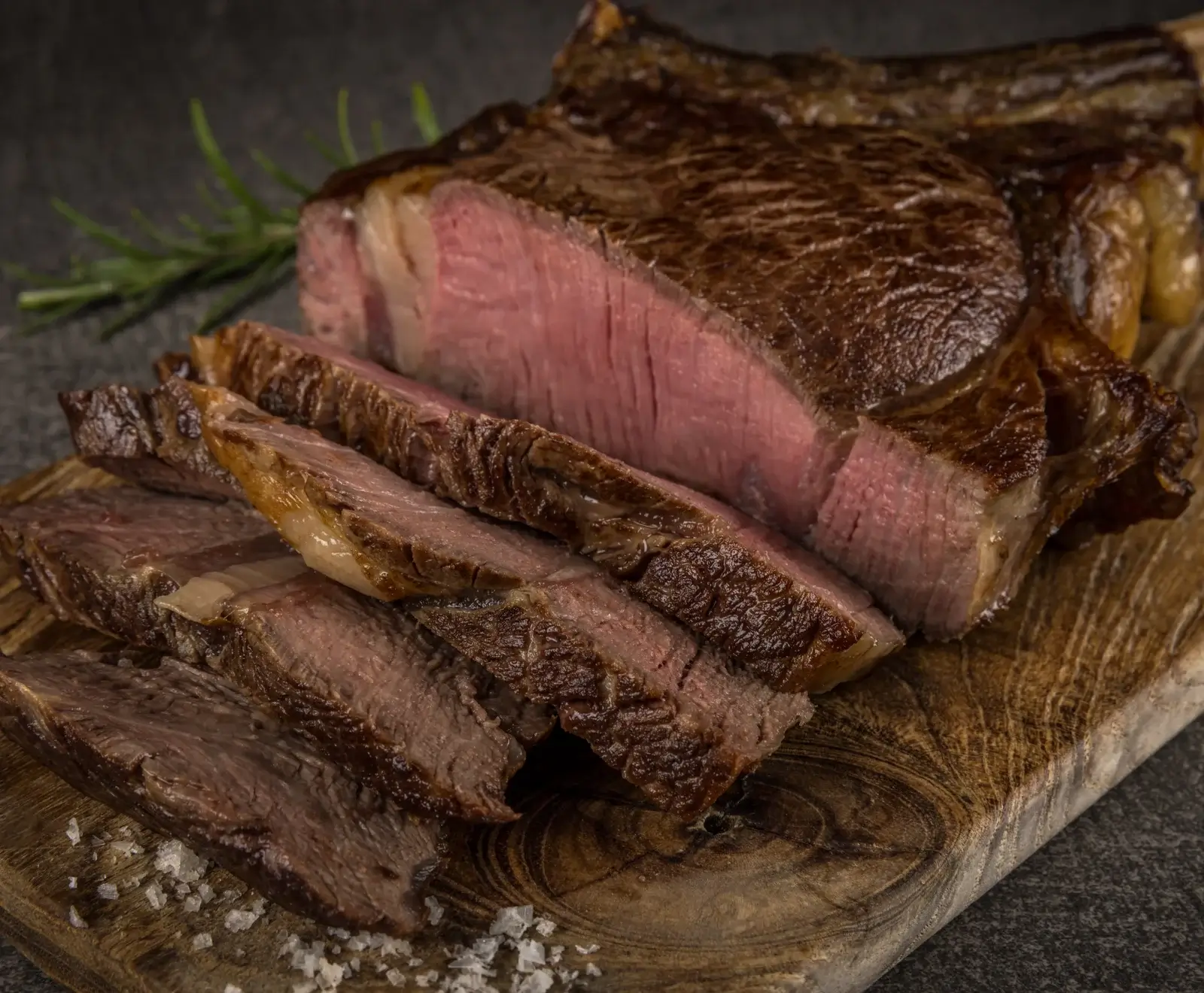 Ein saftiges, medium-rare gegartes Rindersteak, teilweise in dicke Scheiben geschnitten, liegt auf einem rustikalen Holzschneidebrett. Grobes Meersalz und ein Rosmarinzweig sind sichtbar.