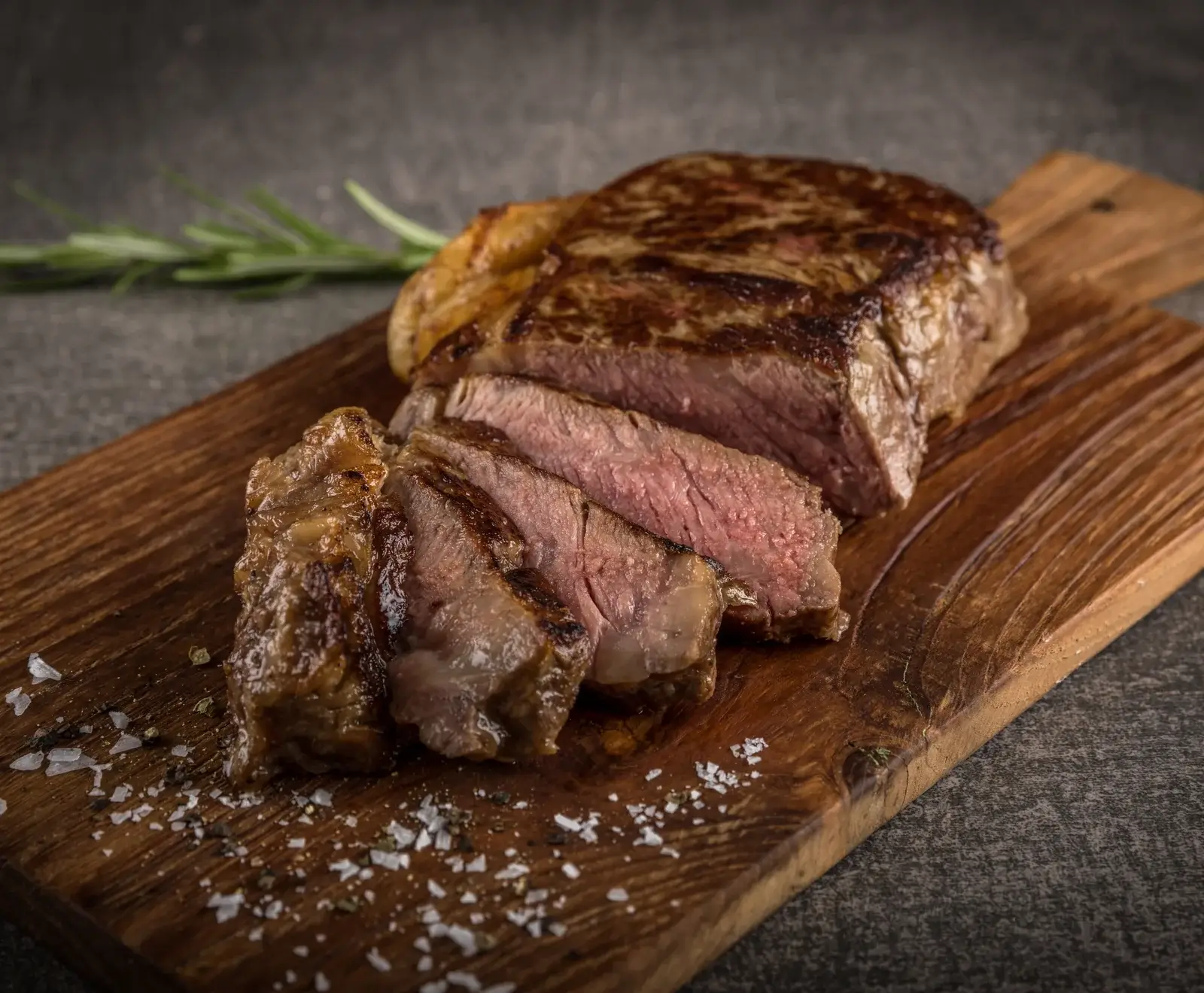 Ein perfekt gebratenes, aufgeschnittenes Steak auf einem Holzbrett, mit grobem Salz und Pfeffer bestreut. Im Hintergrund ist ein Rosmarinzweig zu sehen.