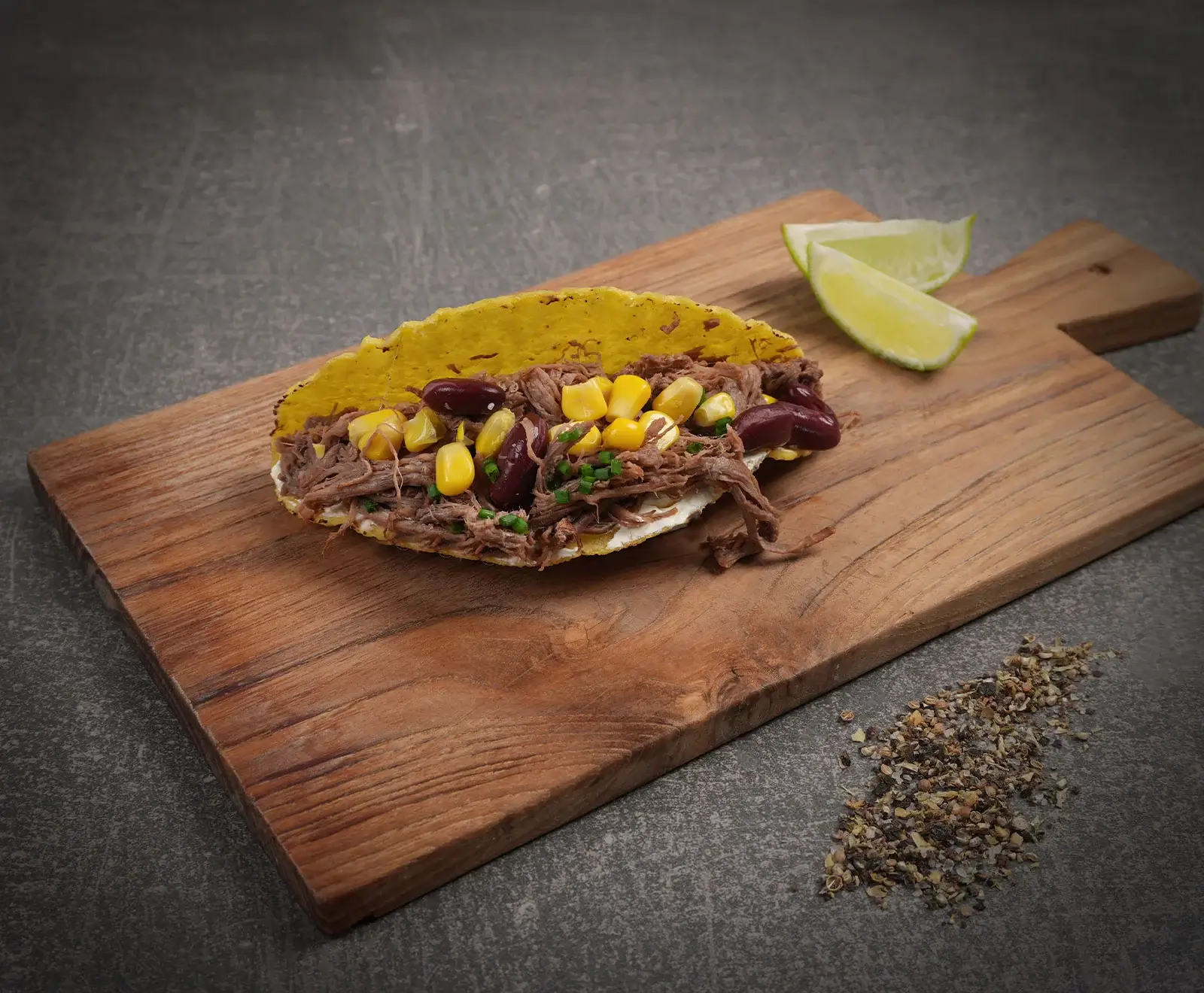 Ein offener Taco mit gelber Maisschale, gefüllt mit gezupftem Fleisch, Mais, Kidneybohnen und Schnittlauch, auf einem Holzbrett mit Limettenspalten und grobem Pfeffer.