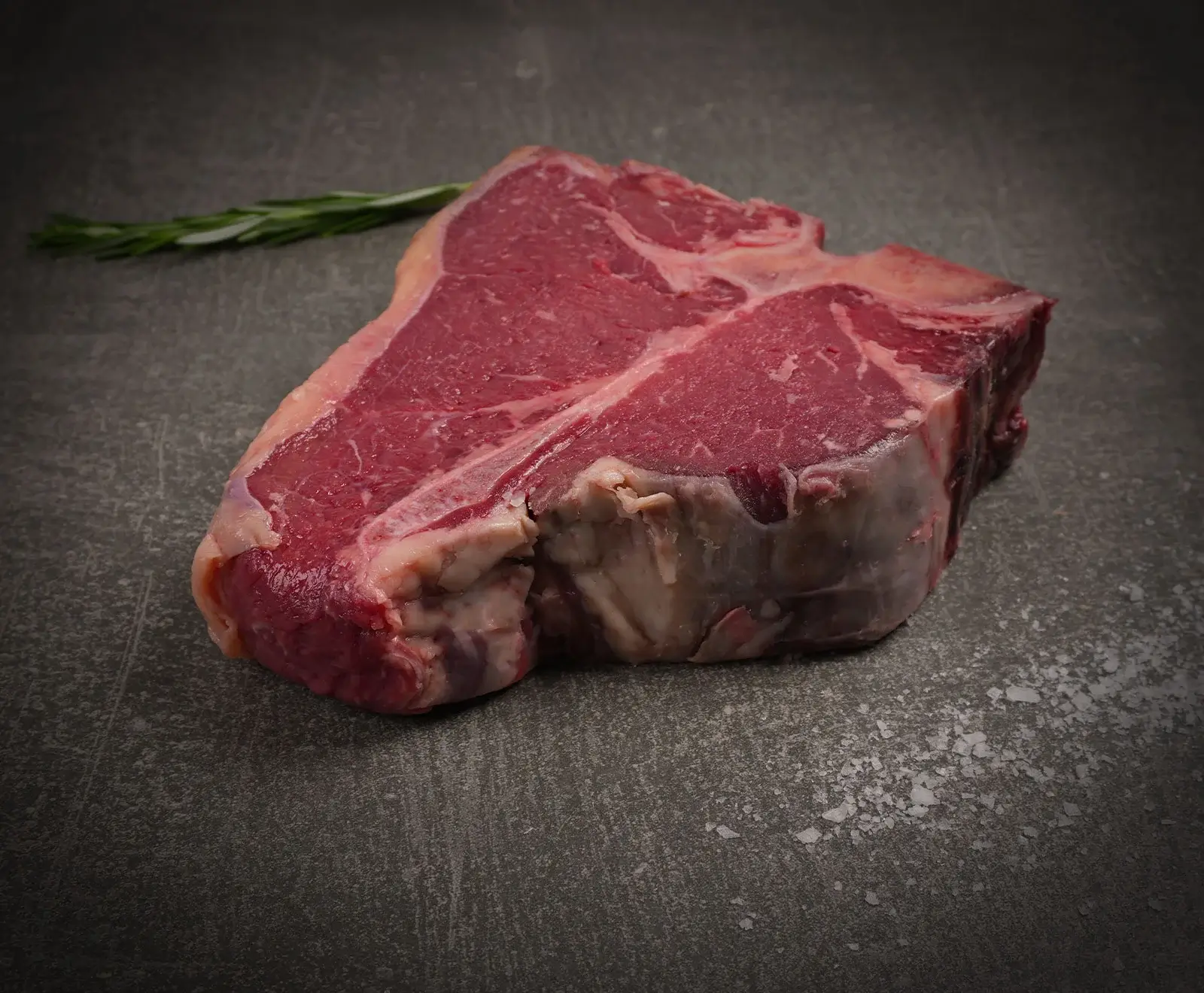 Ein rohes T-Bone-Steak liegt auf einer dunklen, strukturierten Oberfläche. Im oberen linken Hintergrund ist ein Rosmarinzweig zu sehen, während im unteren rechten Vordergrund grobe Salzkristalle verstreut sind.