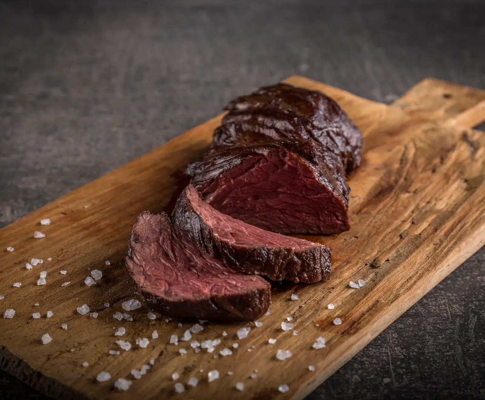 Ein aufgeschnittenes, medium-rare gebratenes Stück Fleisch mit dunkler Kruste liegt auf einem rustikalen Holzbrett. Grobe Meersalzkristalle sind um das Fleisch verstreut. Dunkler, strukturierter Hintergrund.