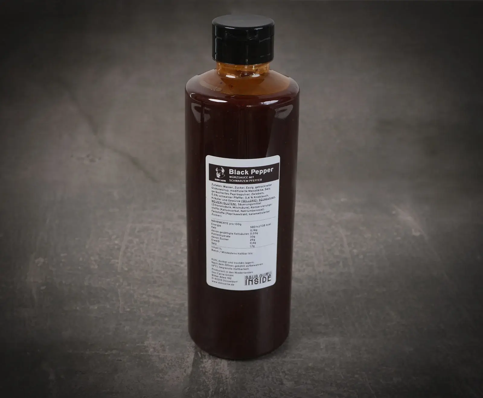 Eine braune Flasche Don Carne Black Pepper Würzsauce mit schwarzem Deckel auf dunklem, strukturiertem Hintergrund. Das weiße Etikett mit Zutaten und Nährwerten ist gut lesbar.