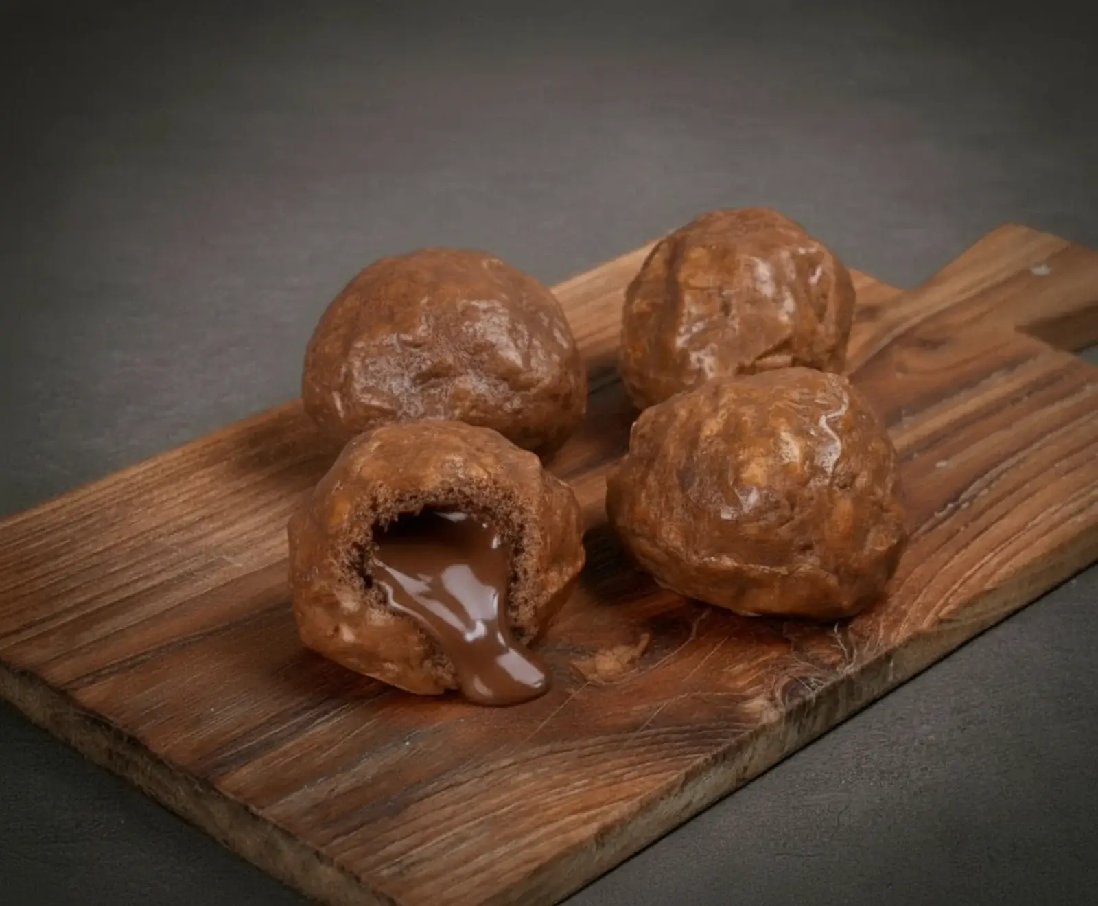 Vier braune, runde Gebäckstücke auf einem Holzbrett. Eines ist geöffnet, mit flüssiger Schokoladenfüllung, die herausläuft. Dunkler Hintergrund.