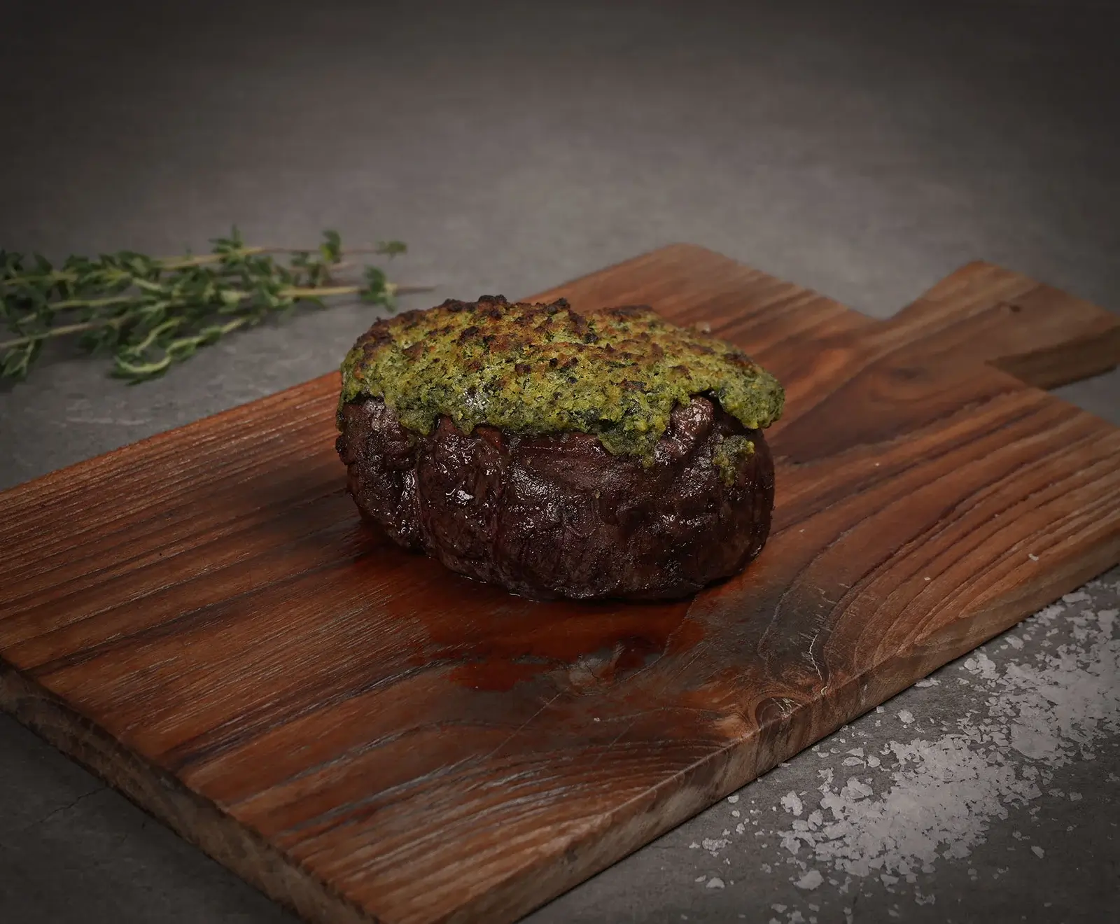 Ein saftiges, dunkelbraunes Steak mit einer dicken grünen Kräuterkruste liegt auf einem rustikalen Holzbrett. Im Hintergrund sind Thymianzweige, im Vordergrund grobes Meersalz auf dunklem Untergrund zu sehen.