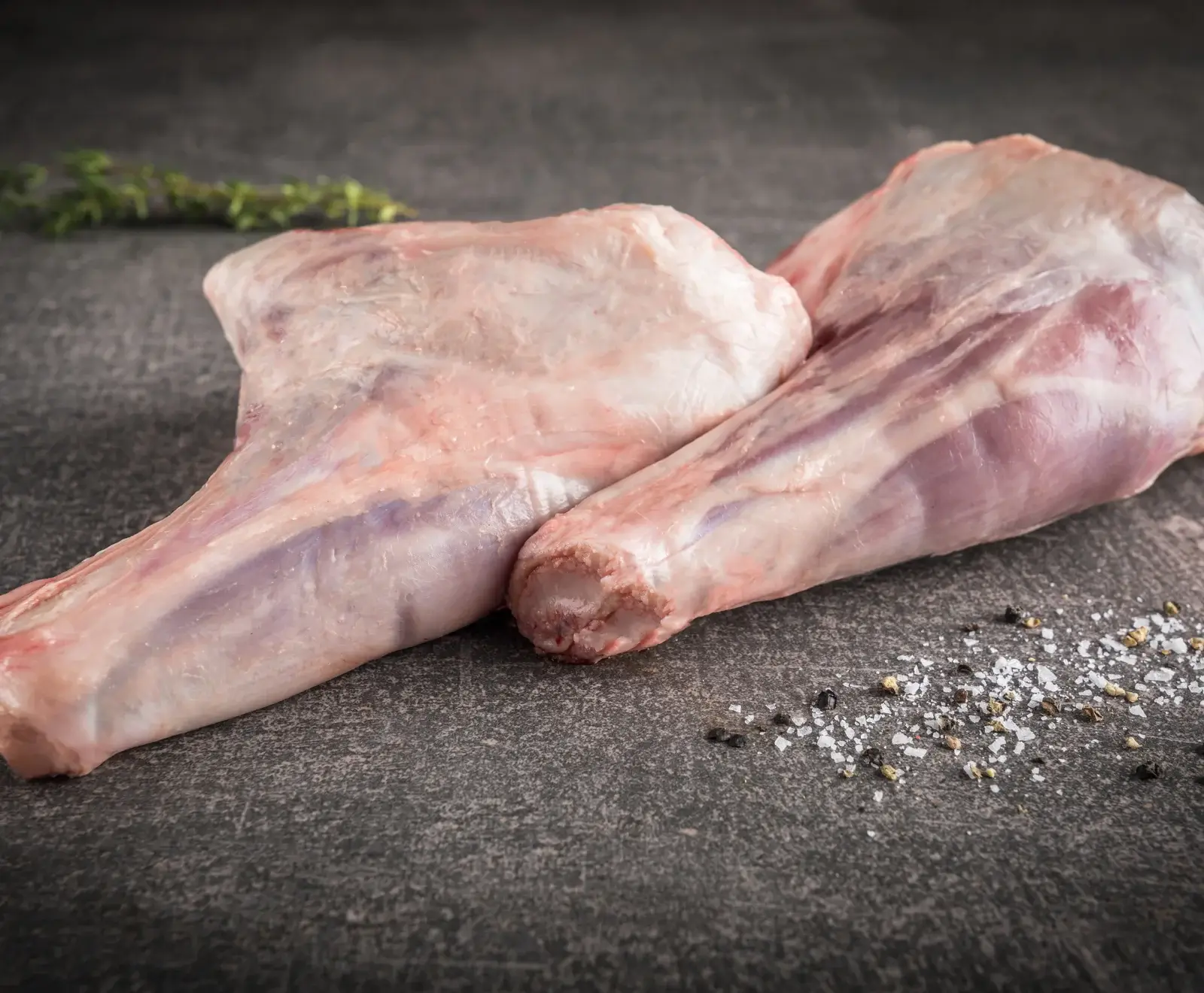 Zwei rohe Lammhaxen liegen auf einer dunklen Steinplatte, daneben ein Zweig Thymian sowie verstreute Salz- und Pfefferkörner. Das Fleisch ist hellrosa mit sichtbaren Knochenenden.