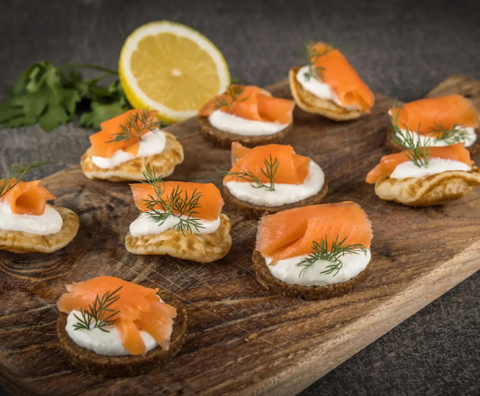 Nahaufnahme von mehreren Lachs-Canapés mit Frischkäse und Dill auf Blinis und dunklem Brot, angerichtet auf einem Holzbrett. Im Hintergrund eine halbe Zitrone und Kräuter.