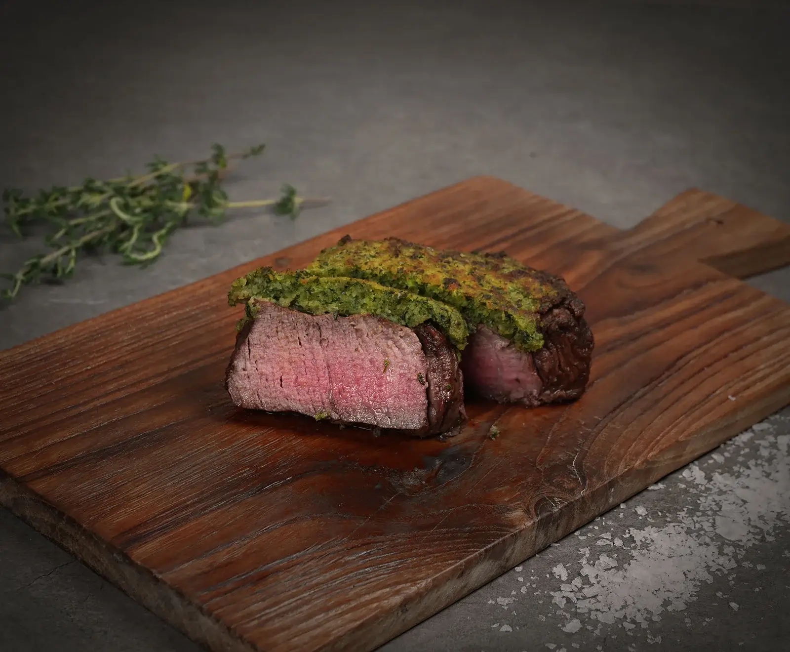 Ein aufgeschnittenes, medium-rare Rinderfilet mit grüner Kräuterkruste auf einem rustikalen Holzbrett. Im Hintergrund liegen Thymianzweige und grobes Meersalz.