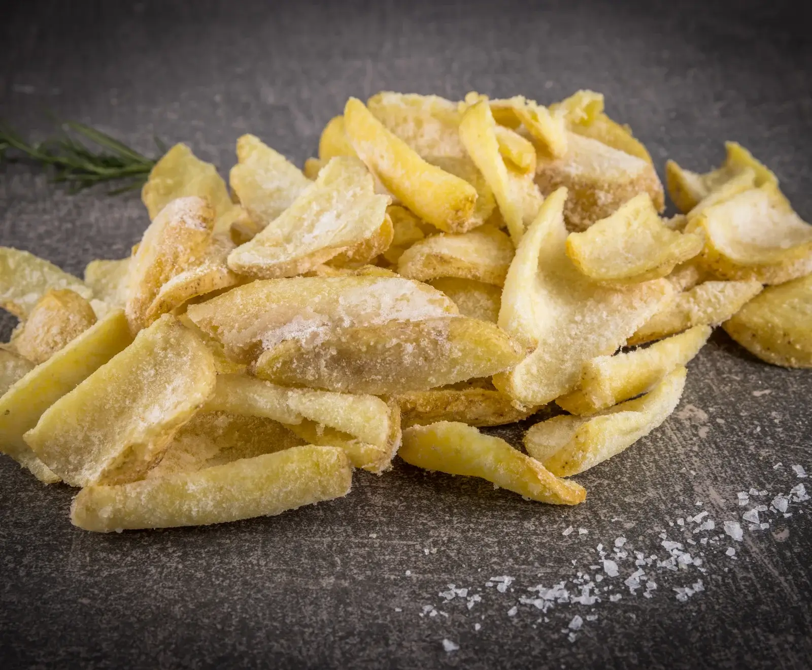 Ein Haufen gefrorener, mit Eiskristallen bedeckter Kartoffelspalten liegt auf einer dunklen, strukturierten Oberfläche. Im Hintergrund ist ein Rosmarinzweig zu sehen, im Vordergrund verstreute Salzkörner.