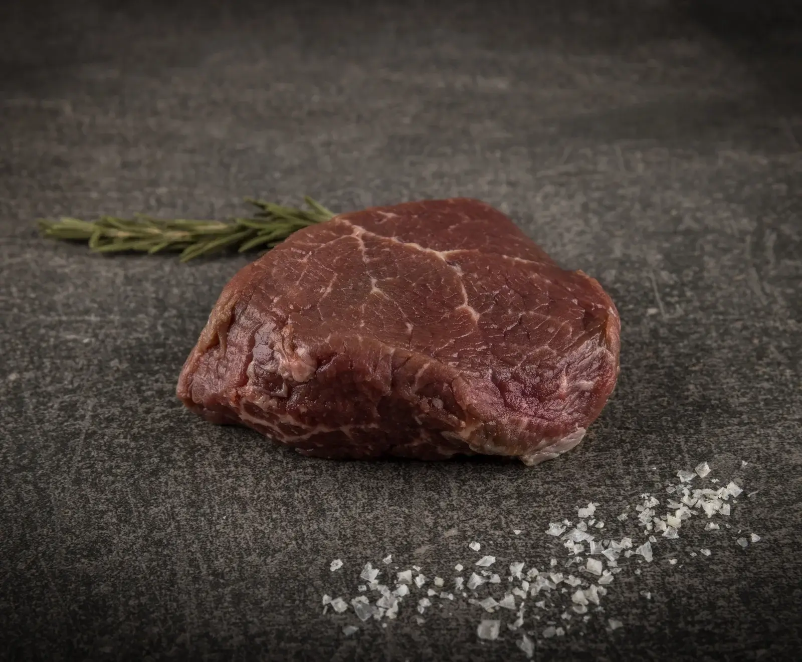Nahaufnahme eines rohen Rinderfilets auf dunkler Steinplatte, begleitet von einem Rosmarinzweig und groben Meersalzkristallen.