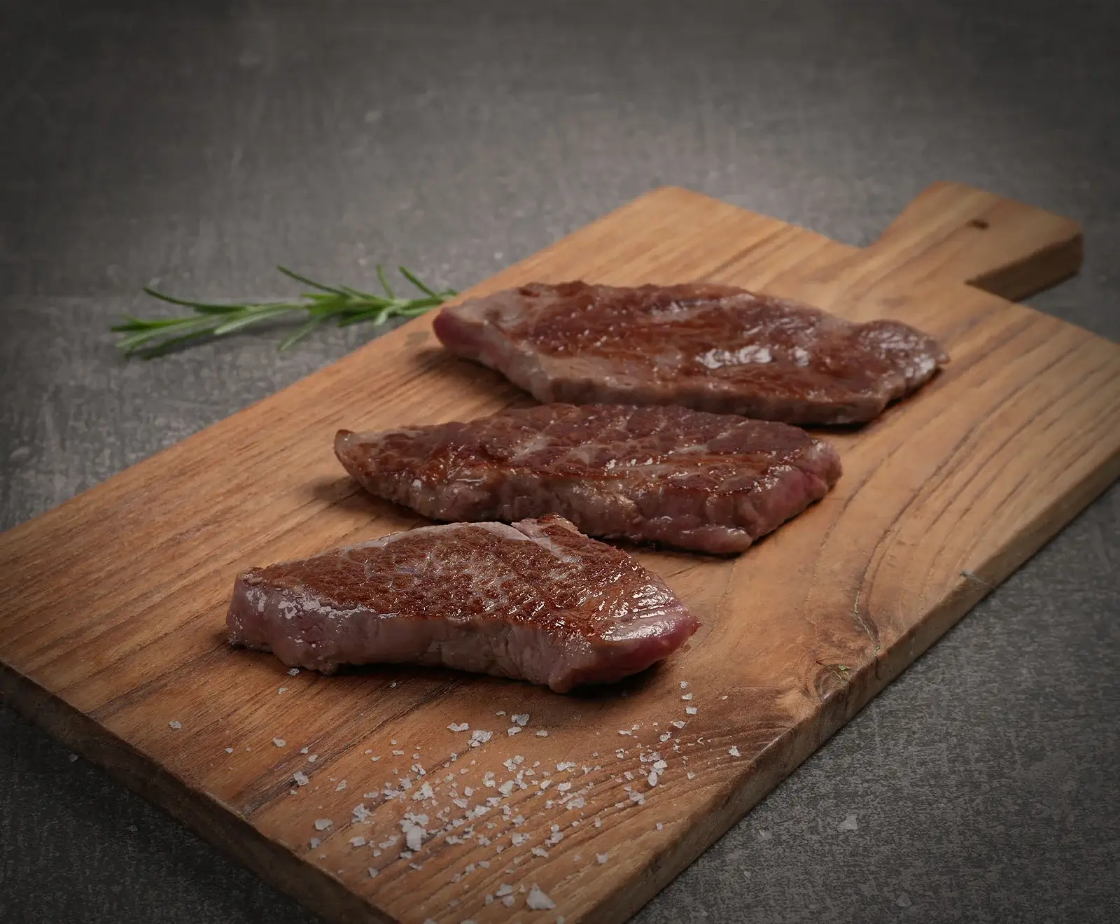 Drei gebratene Steaks mit brauner Kruste und rosa Kern liegen diagonal auf einem Holzschneidebrett. Ein Rosmarinzweig und grobes Salz sind ebenfalls auf dem Brett zu sehen. Der Hintergrund ist dunkelgrau.