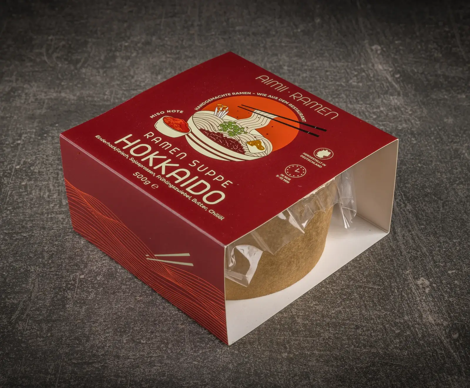 Eine rote, teilweise geöffnete Verpackung von Aimi Ramen Suppe Hokkaido auf dunklem Untergrund. Sie zeigt eine Ramen-Illustration und Details wie Miso, Rinderhackfleisch und 5-10 Min. Zubereitungszeit.