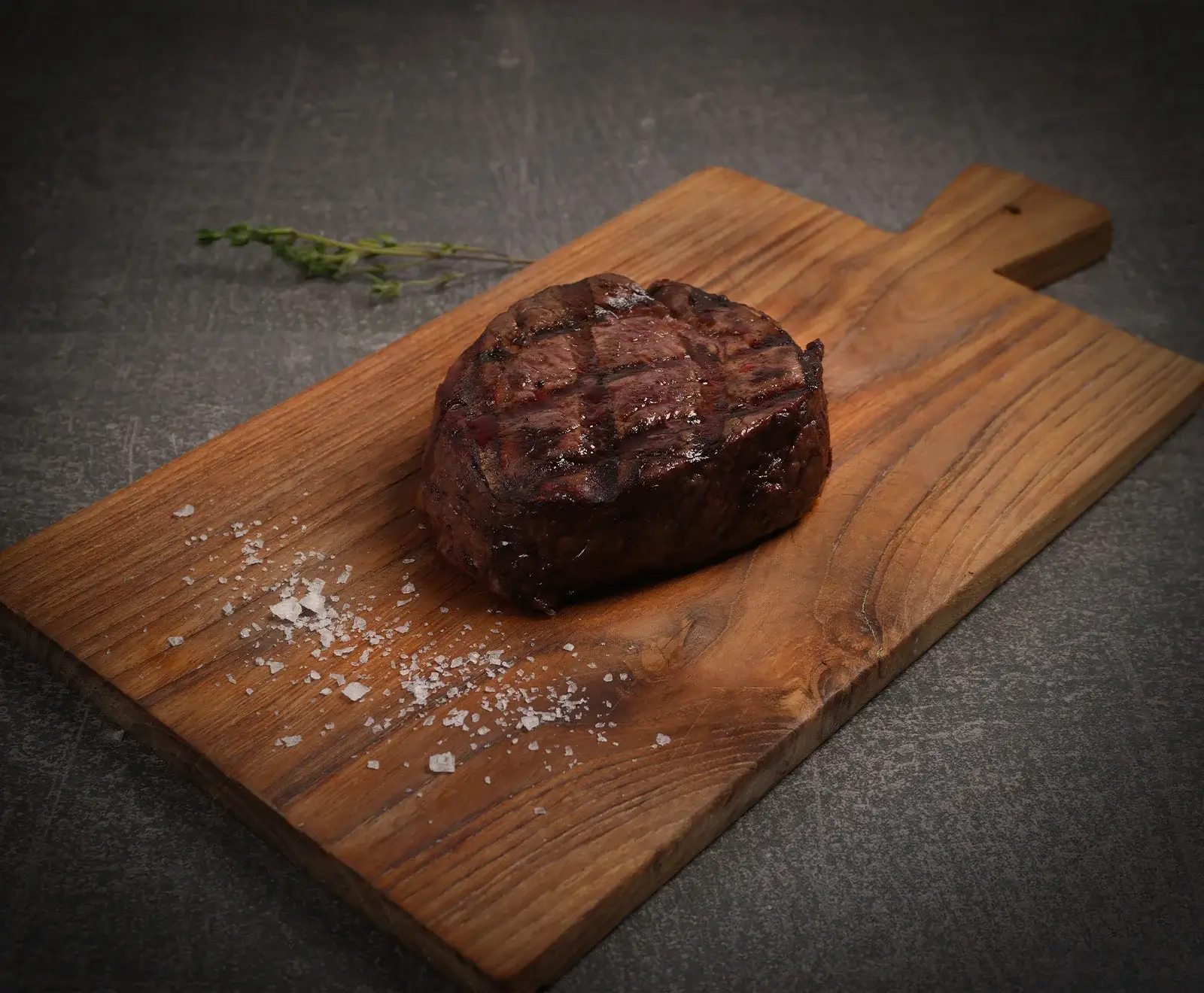Ein perfekt gegrilltes, saftiges Steak mit deutlichen Grillstreifen liegt auf einem rustikalen Holzschneidebrett. Grobes Meersalz und ein Thymianzweig sind daneben auf dunklem Hintergrund platziert.
