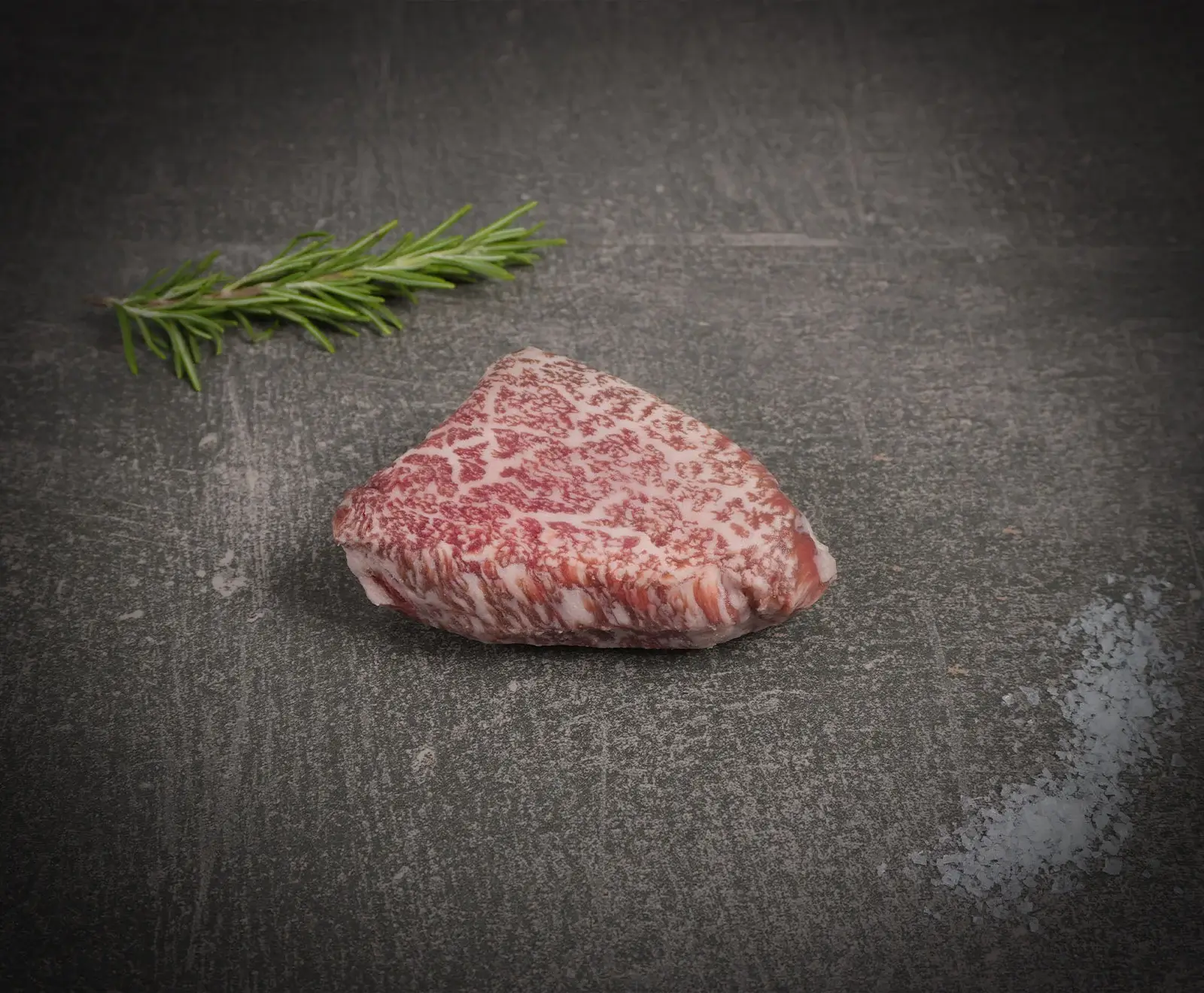 Ein rohes, stark marmoriertes Rindersteak (vermutlich Wagyu) liegt auf einer dunklen Oberfläche, begleitet von einem Rosmarinzweig und grobem Meersalz.