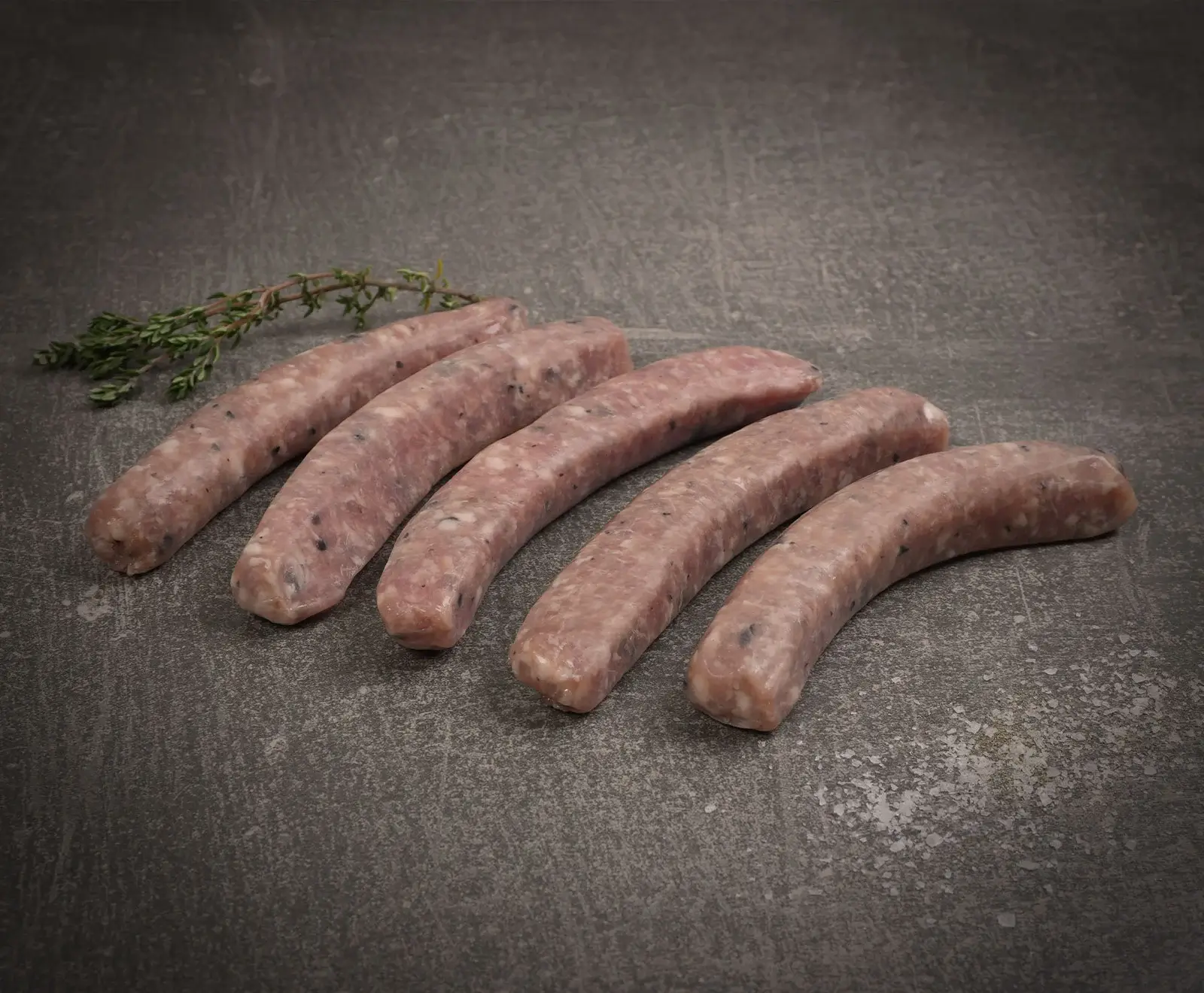 Fünf rohe, hellrosa Würste mit dunklen Sprenkeln liegen leicht gekrümmt auf einer dunklen, strukturierten Oberfläche. Ein frischer Thymianzweig liegt links oben.