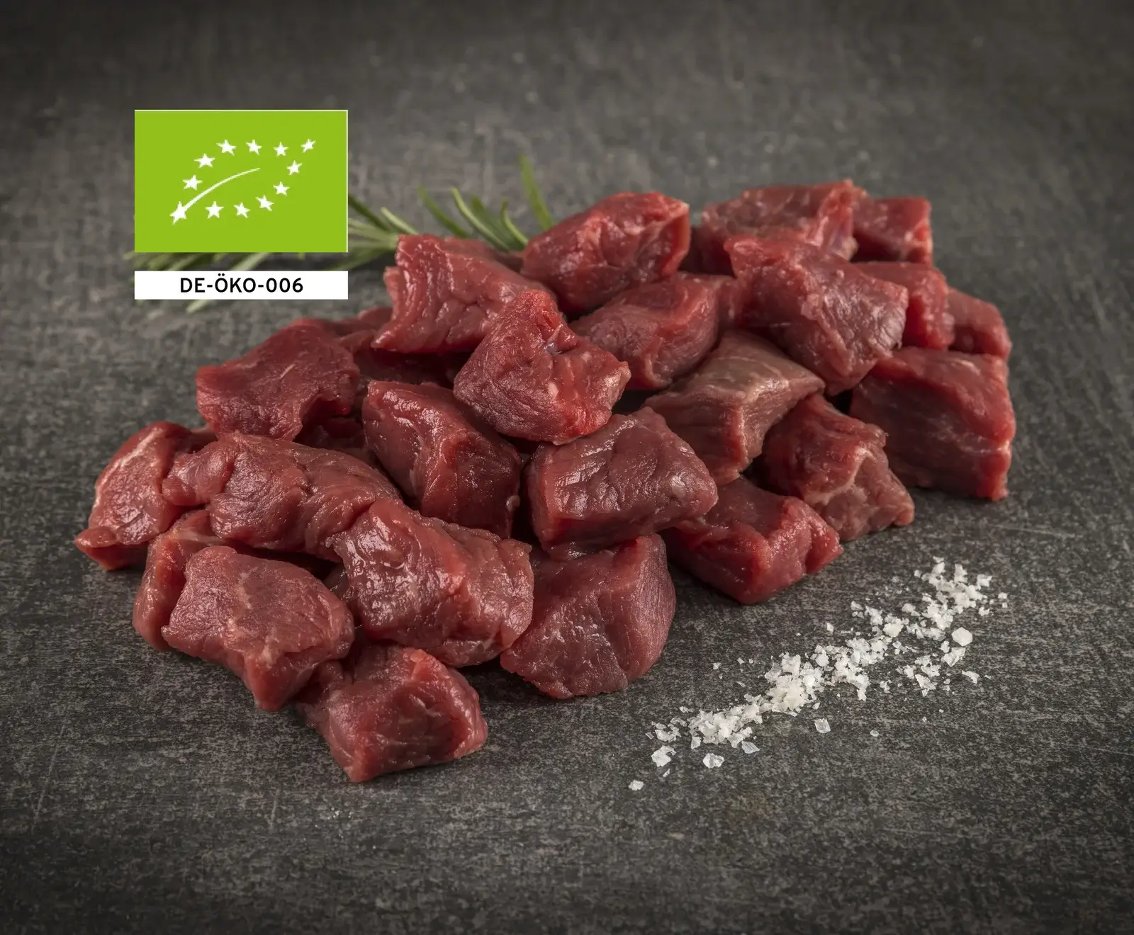 Ein Haufen rohes, gewürfeltes Rindfleisch auf dunklem Untergrund, mit Rosmarin, Meersalz und dem EU-Bio-Siegel DE-ÖKO-006 in der oberen linken Ecke.