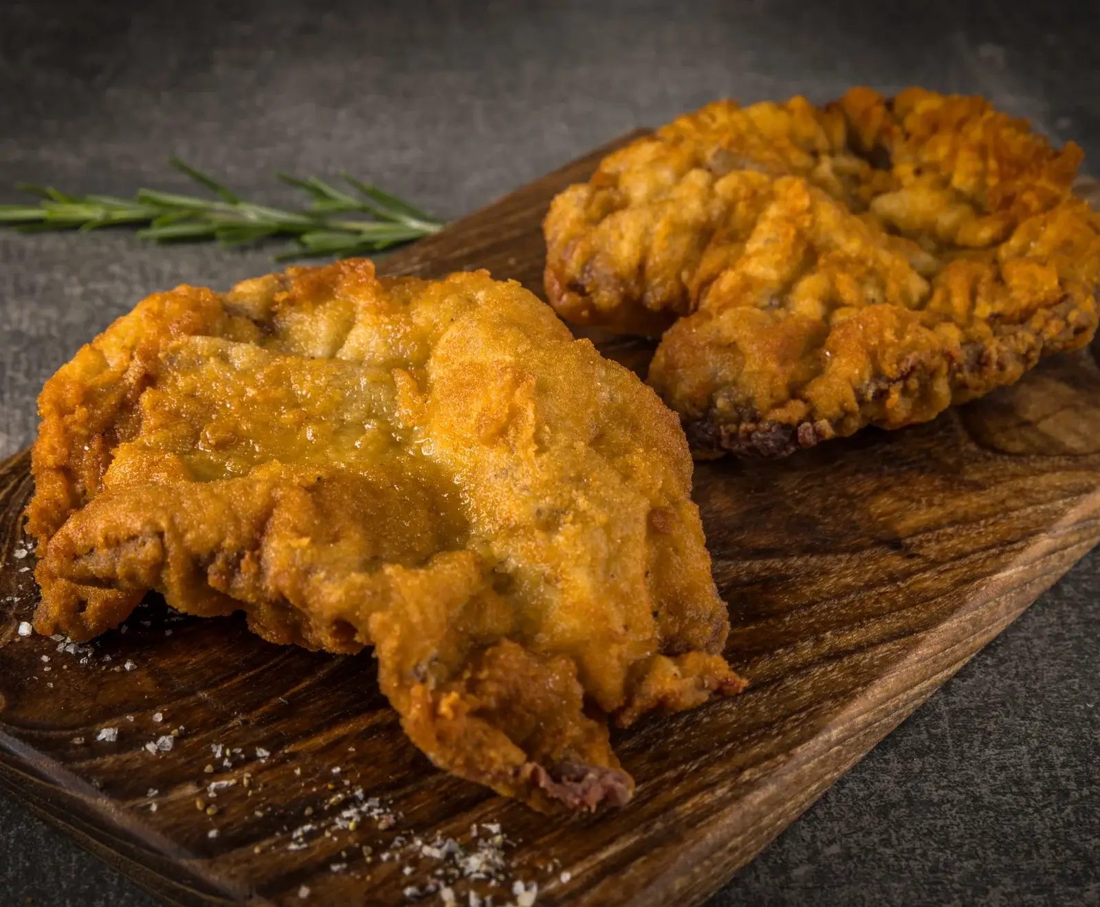 Zwei frisch gebratene, goldbraune Schnitzel auf einem dunklen Holzschneidebrett, mit Rosmarin und grobem Salz bestreut. Der Hintergrund ist dunkelgrau.