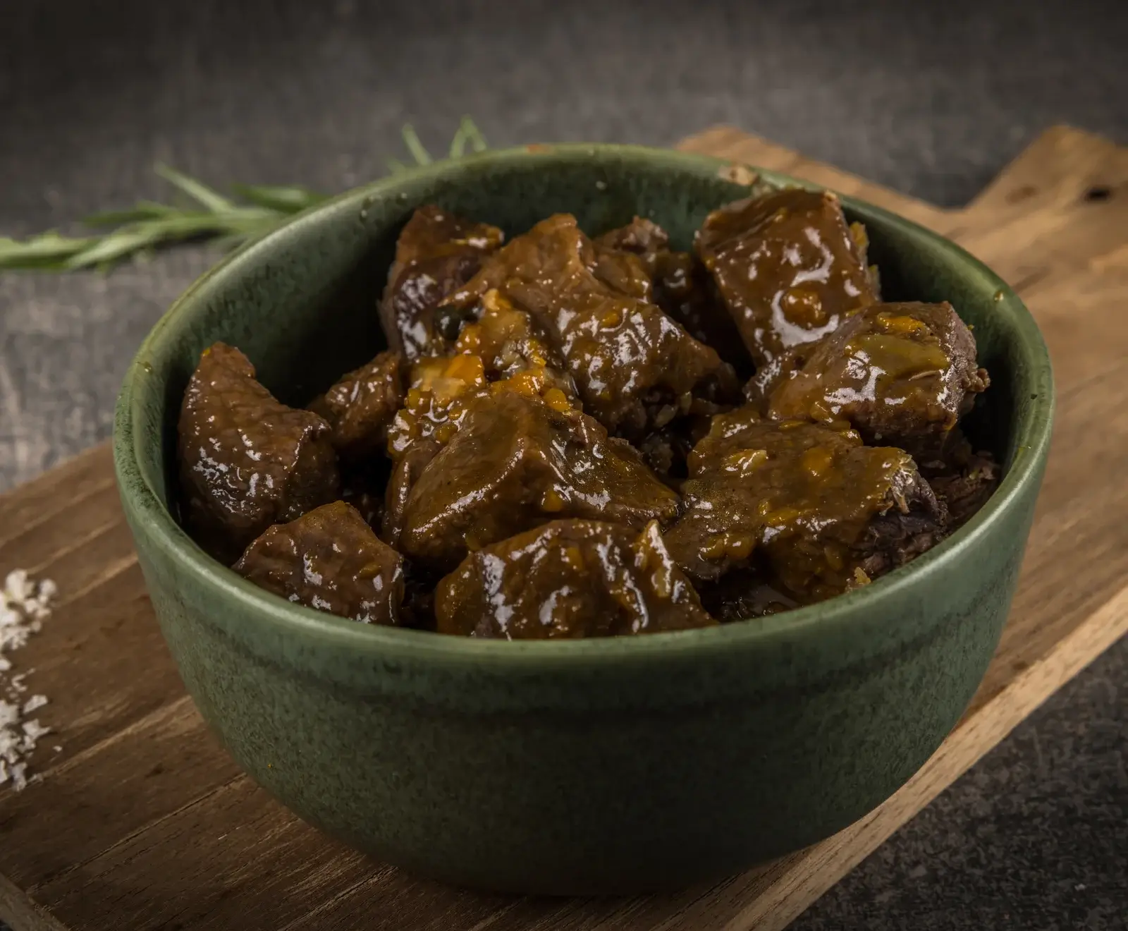 Eine dunkelgrüne Keramikschale, gefüllt mit glänzendem, braunem Gulasch oder Schmorgericht aus zarten Fleischstücken in einer reichhaltigen Soße. Angerichtet auf einem Holzbrett mit Rosmarin und Salzflocken auf dunklem Hintergrund.