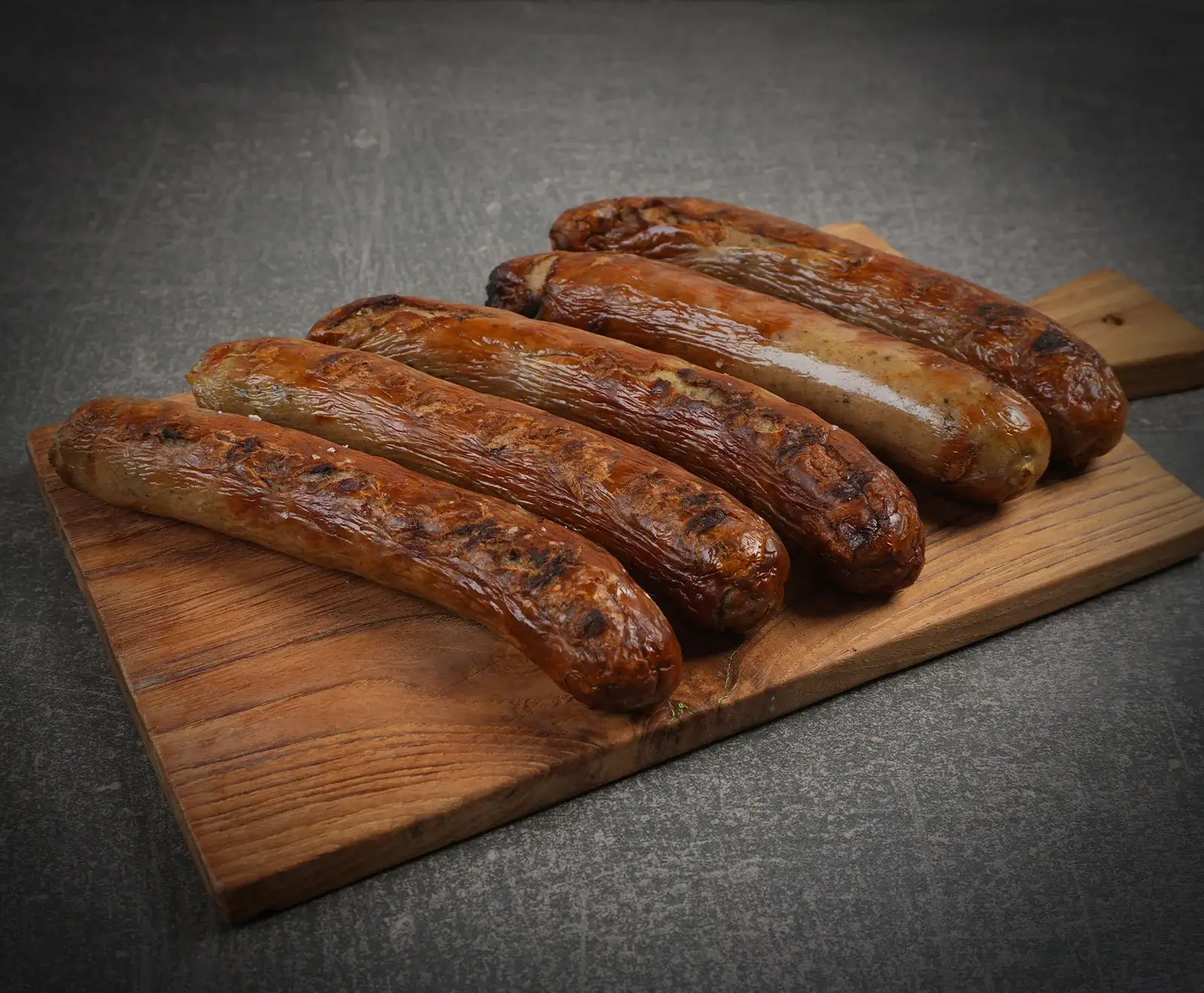 Sechs goldbraun gegrillte Bratwürste mit deutlichen Grillspuren liegen diagonal auf einem rustikalen Holzbrett vor einem dunklen, strukturierten Hintergrund.