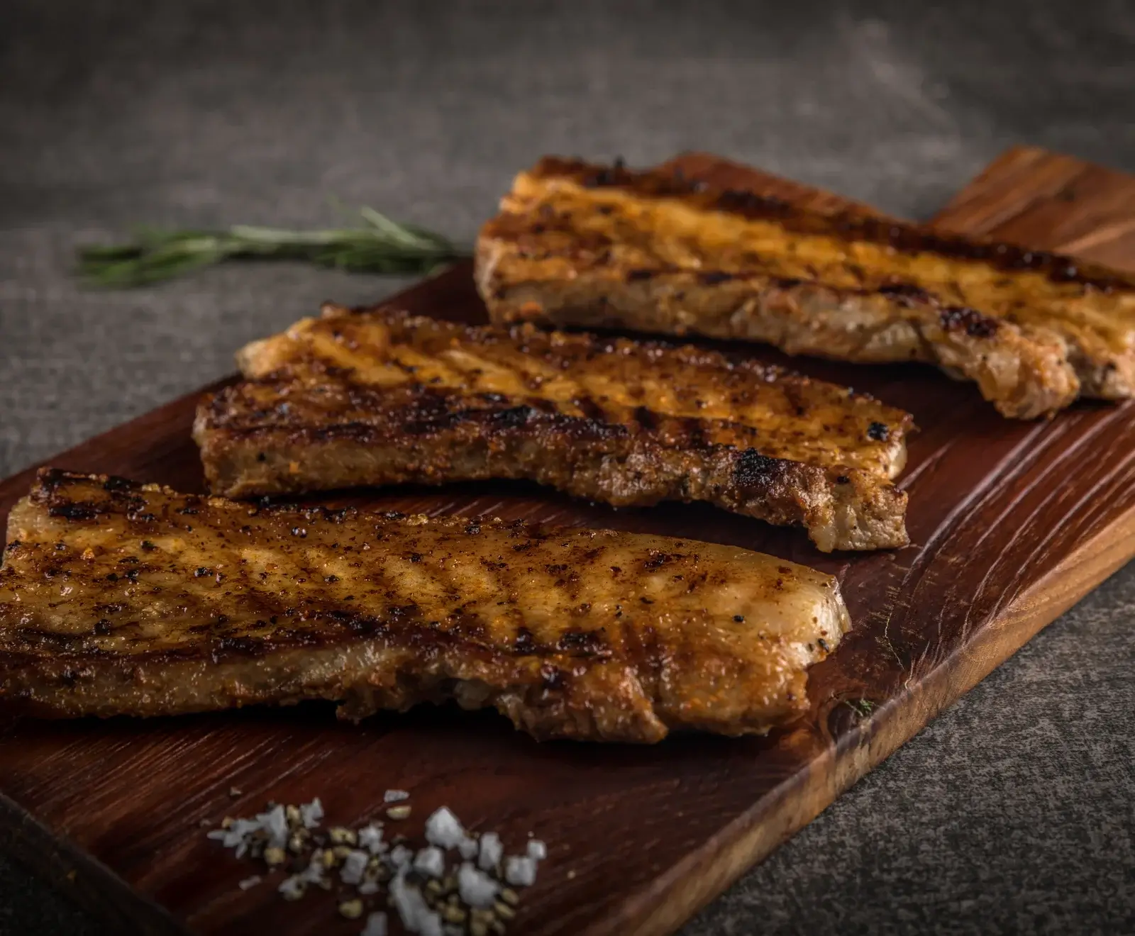 Drei knusprig gegrillte Schweinebauchscheiben liegen auf einem dunklen Holzschneidebrett. Im Vordergrund sind grobes Salz und Pfefferkörner zu sehen, im Hintergrund ein Rosmarinzweig.