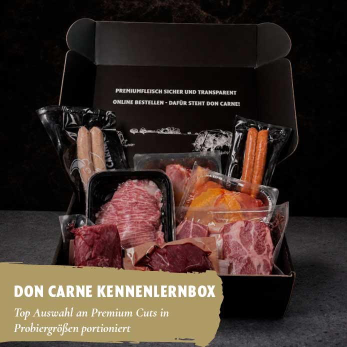 Eine offene schwarze Box, gefüllt mit verschiedenen vakuumverpackten Premium-Fleischstücken wie Steaks und Würsten. Der Deckel enthält Text. Unten links steht "DON CARNE KENNLERNBOX Top Auswahl an Premium Cuts in Probiergrößen portioniert".
