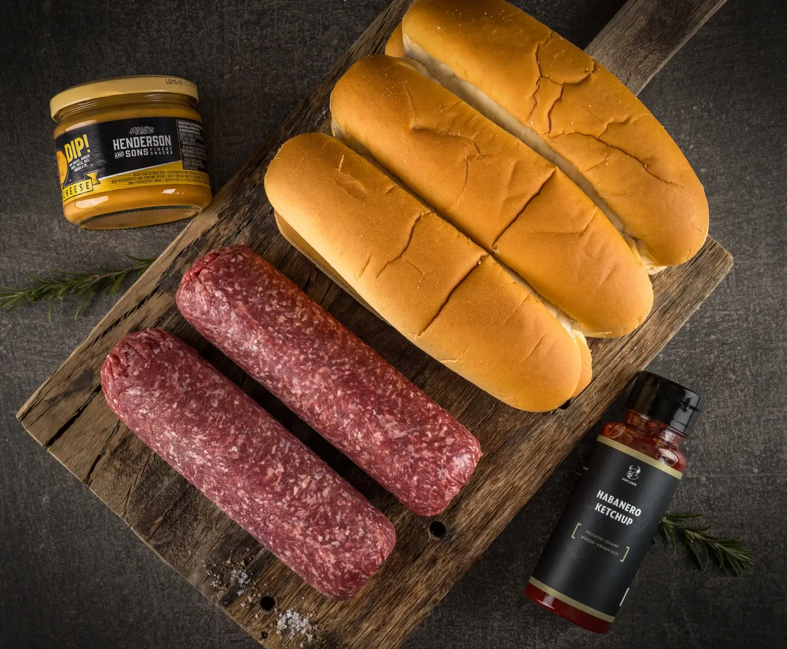 Zwei rohe Würste, drei Hotdog-Brötchen, ein Glas Käse-Dip und eine Flasche Habanero-Ketchup auf einem rustikalen Holzbrett, bereit für die Zubereitung von Hot Dogs.