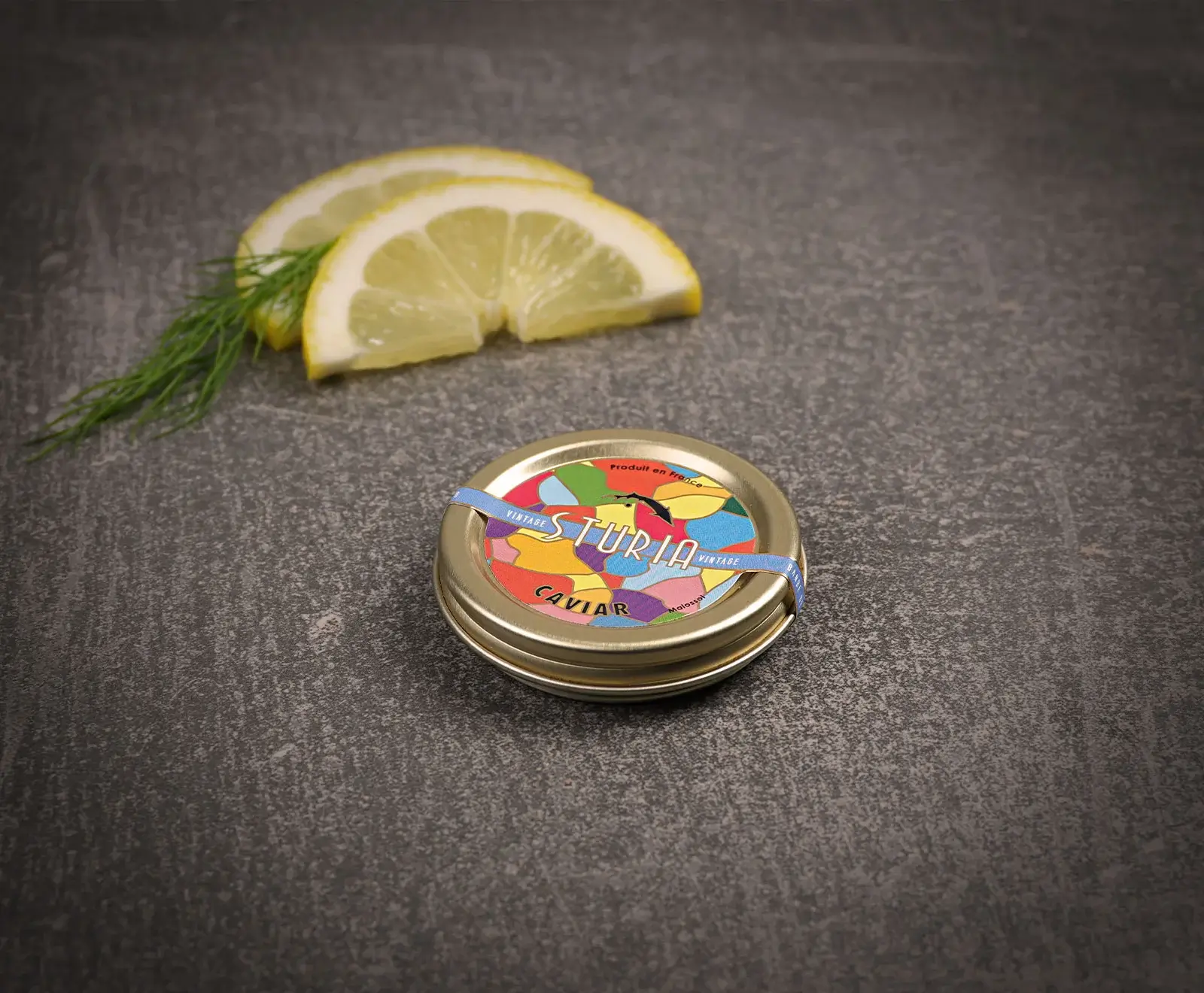 Eine kleine, goldene Dose Sturia Kaviar mit buntem Deckel, daneben zwei Zitronenscheiben und ein Dillzweig, auf dunklem strukturiertem Untergrund.