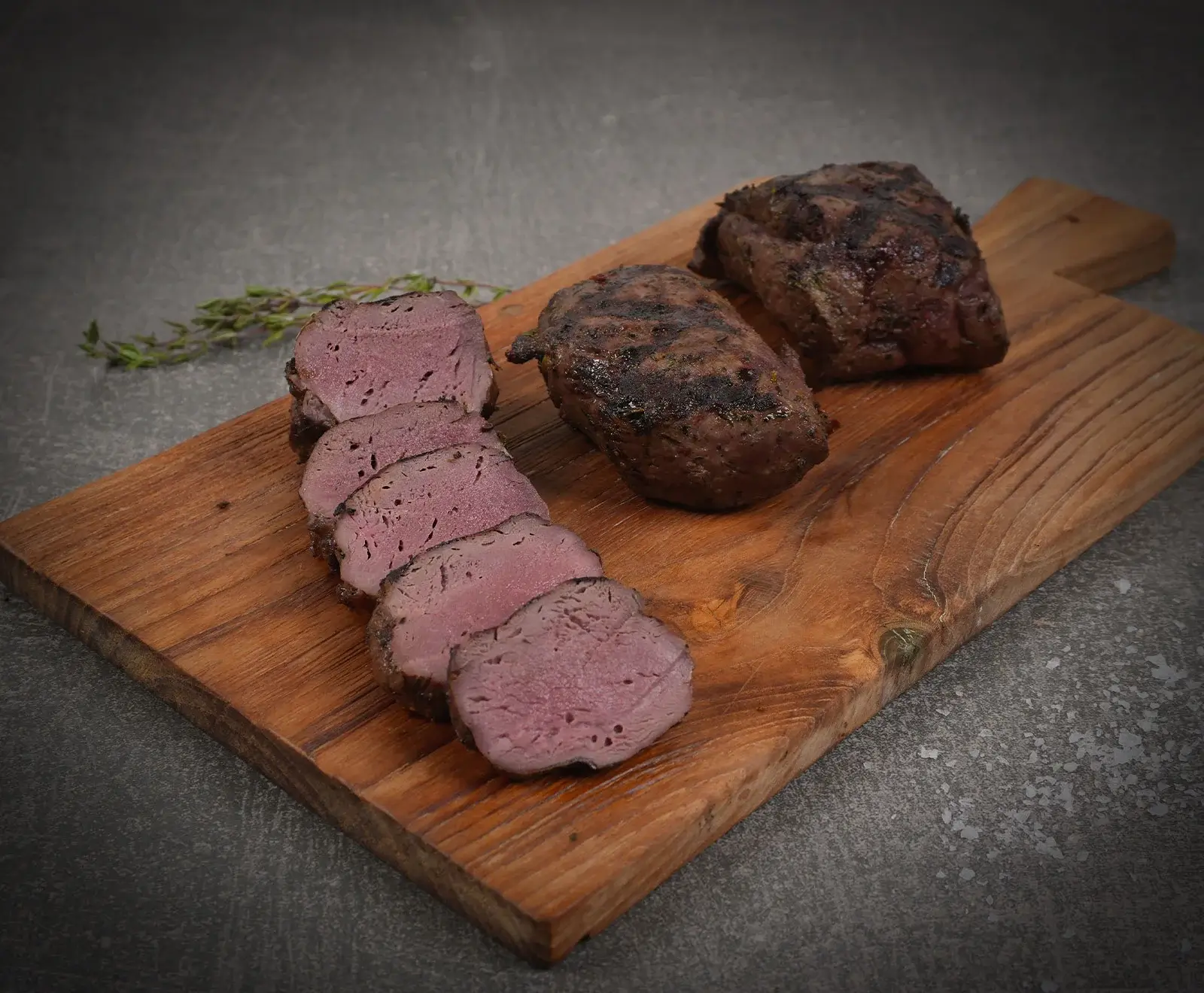 Zwei gegrillte Fleischstücke auf einem Holzschneidebrett. Eines ist in Scheiben geschnitten und zeigt eine rosa Mitte. Ein Thymianzweig liegt daneben.