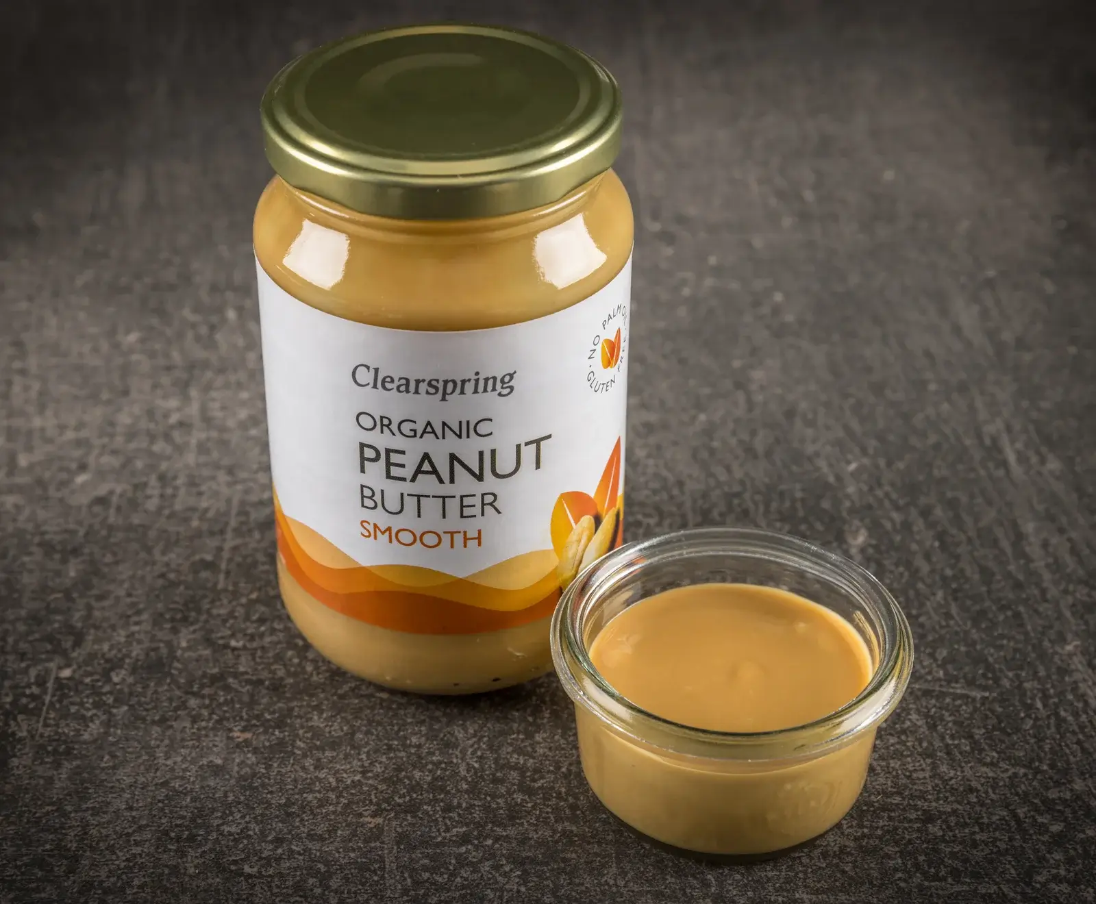 Ein Glas Clearspring Bio Erdnussbutter Smooth mit goldenem Deckel steht neben einer kleinen Glasschale, die ebenfalls glatte Erdnussbutter enthält, auf dunklem, strukturiertem Untergrund.