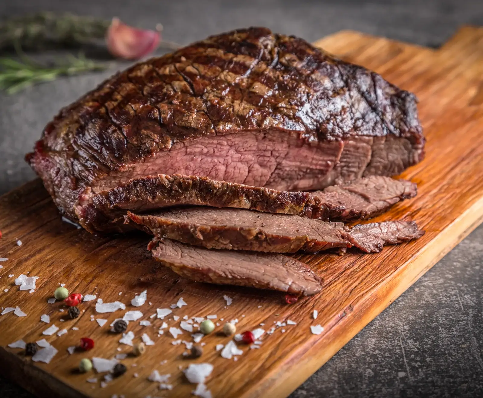 Ein saftiges, gebratenes Stück Rindfleisch, teilweise in dünne Scheiben geschnitten, liegt auf einem Holzschneidebrett. Die Kruste ist dunkelbraun, das Innere rosa. Grobes Meersalz und bunte Pfefferkörner sind verstreut.
