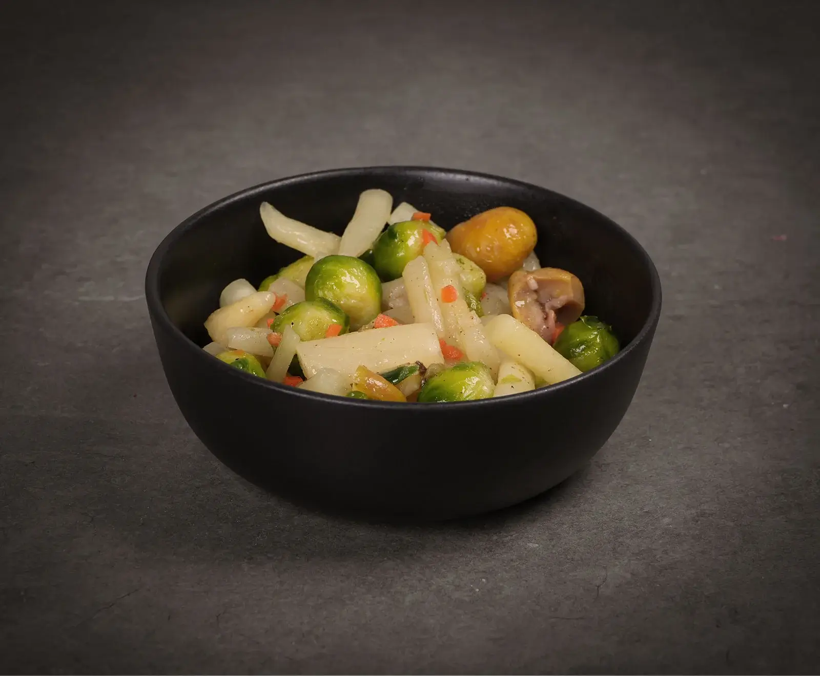 Eine schwarze Schale gefüllt mit einem bunten Gemüsemix. Deutlich sichtbar sind hellgrüner Rosenkohl, weiße Spargelstücke, kleine orangefarbene Karottenwürfel und bräunliche Maronen. Die Schale steht auf einer dunkelgrauen Oberfläche.