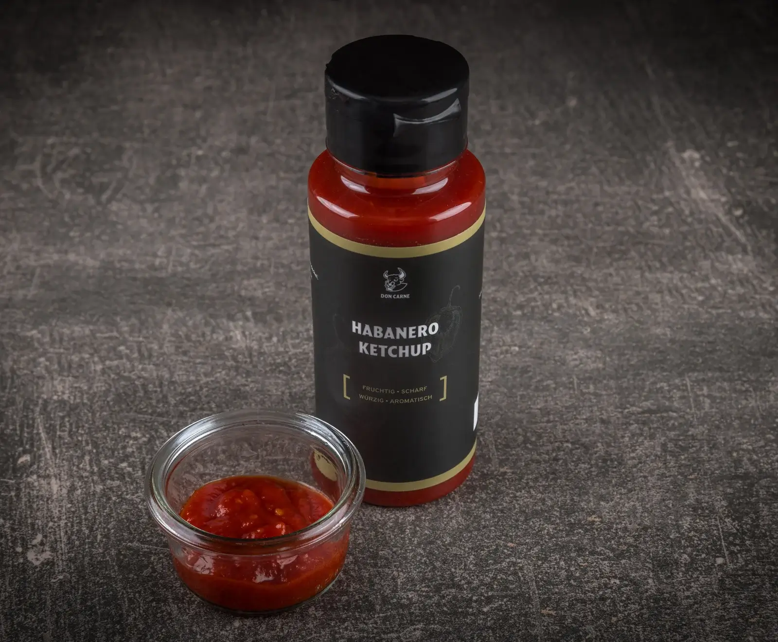 Eine schwarze Flasche Don Carne Habanero Ketchup mit rotem Inhalt und schwarzem Deckel steht auf dunklem Untergrund. Daneben eine kleine Glasschale mit dem roten, stückigen Ketchup.