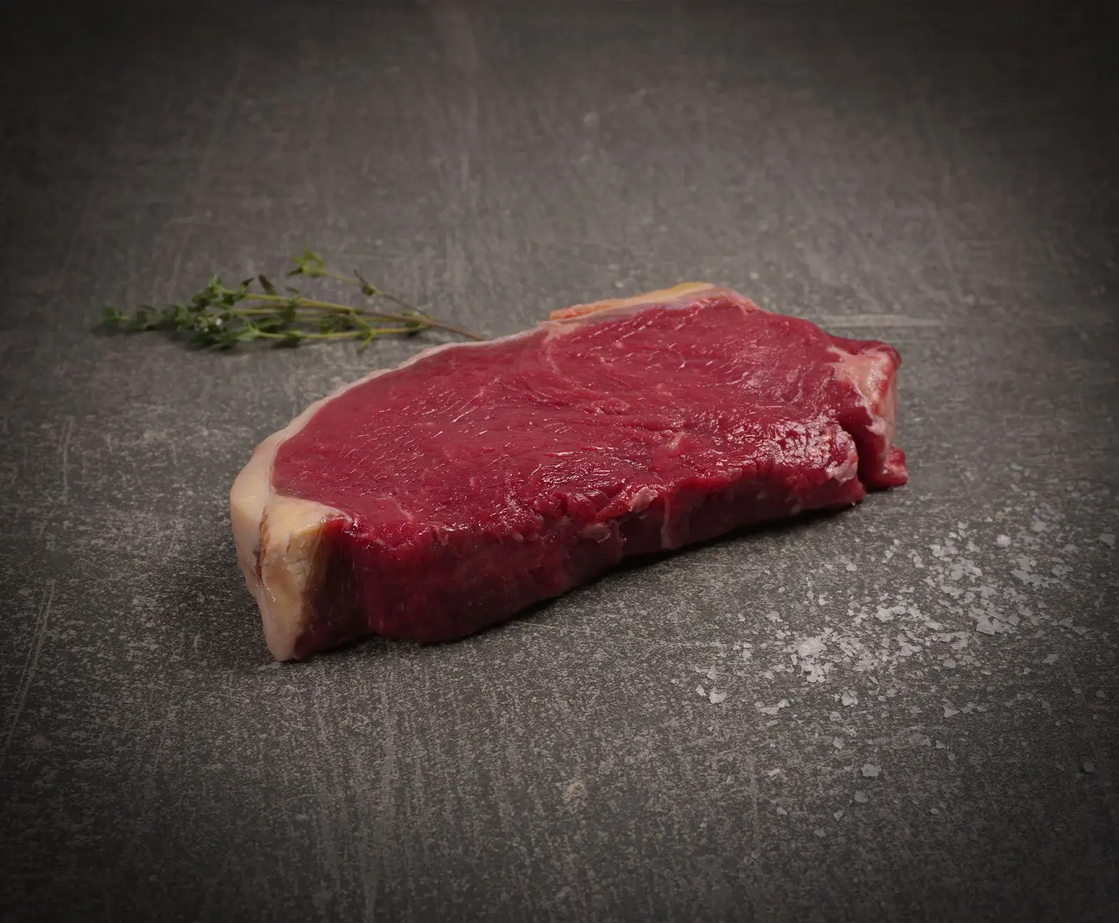 Ein rohes, rotes Steak mit Fettrand liegt auf einer dunklen Steinplatte. Links oben befindet sich ein Zweig frischer Thymian, rechts unten grobes Meersalz.