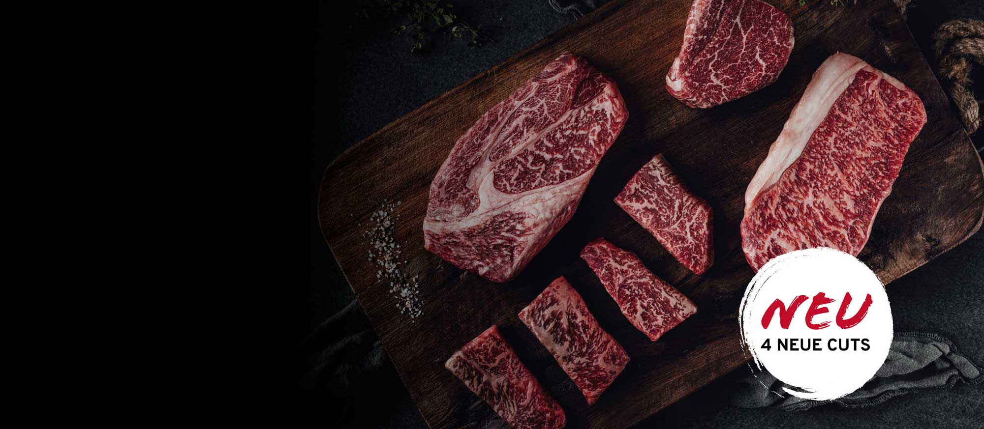 Verschiedene rohe, stark marmorierte Fleischstücke auf einem dunklen Holzbrett mit grobem Salz. Im Vordergrund ein weißes Logo mit rotem Text: NEU, 4 NEUE CUTS.