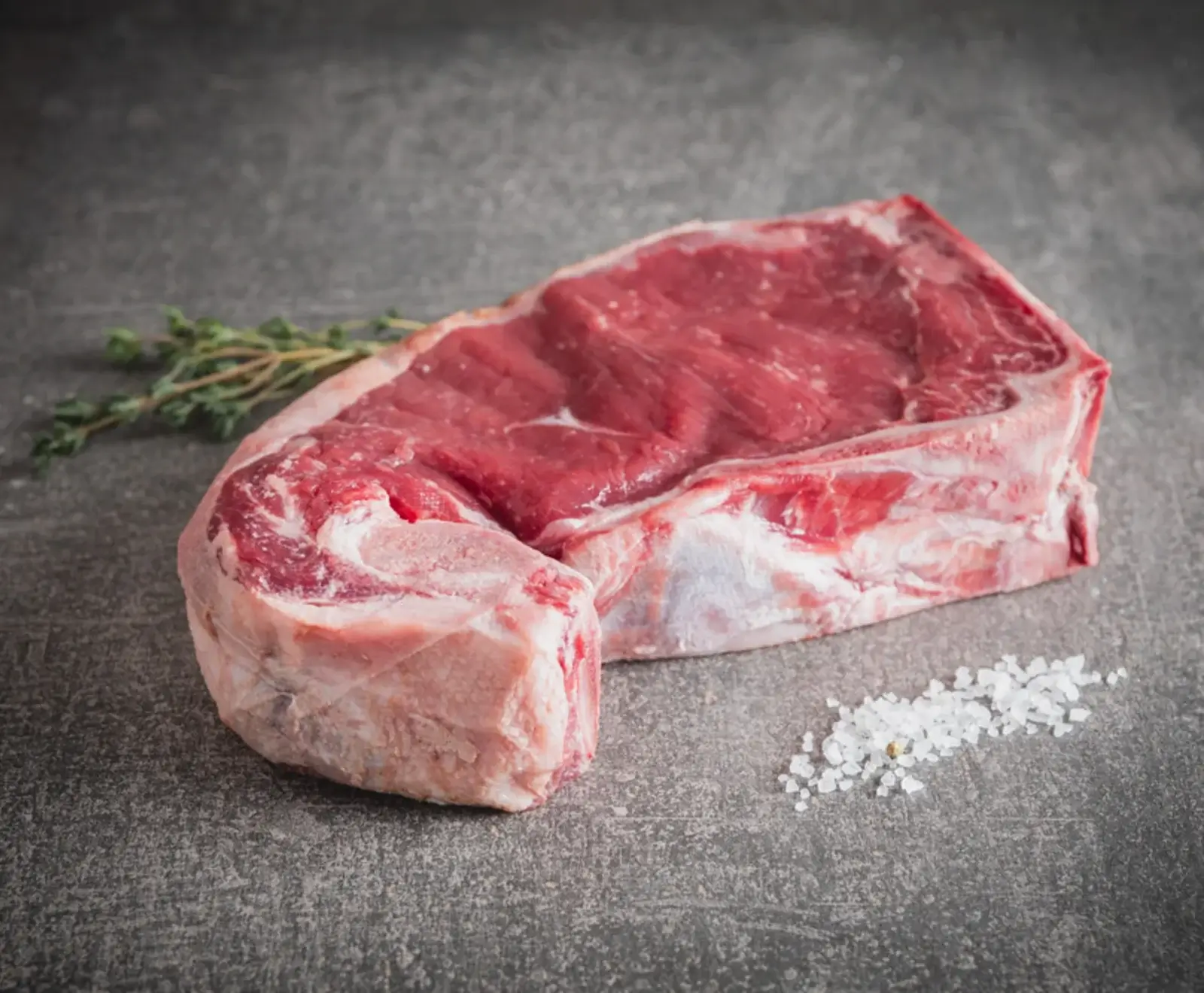Ein rohes Stück Rindersteak mit Knochen und Fettrand liegt auf einer dunklen, strukturierten Oberfläche. Daneben sind ein frischer Thymianzweig und grobes Meersalz zu sehen.