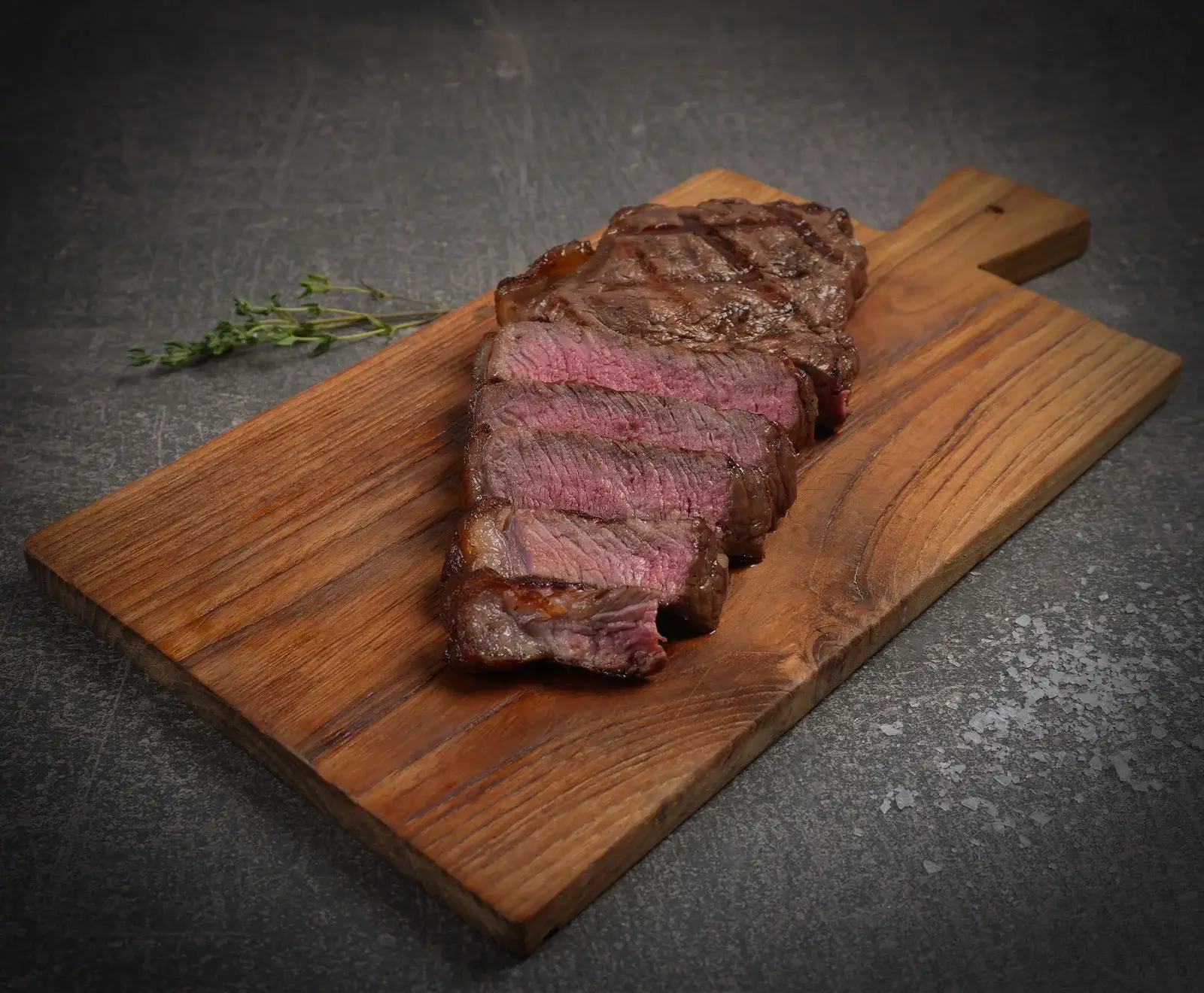 Geschnittenes, medium-rare Steak auf einem Holzschneidebrett mit einem Thymianzweig auf dunklem Hintergrund.