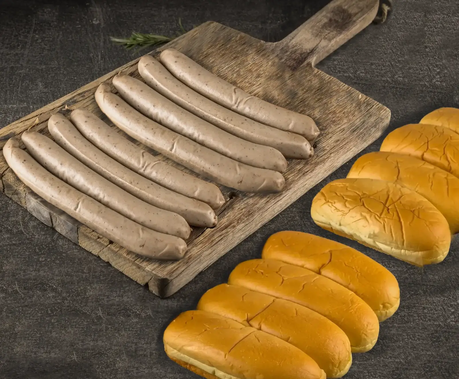 Acht rohe, helle Würste liegen auf einem rustikalen Holzbrett, daneben acht goldbraune Hotdog-Brötchen auf dunklem Untergrund. Ein Rosmarinzweig ist im Hintergrund.