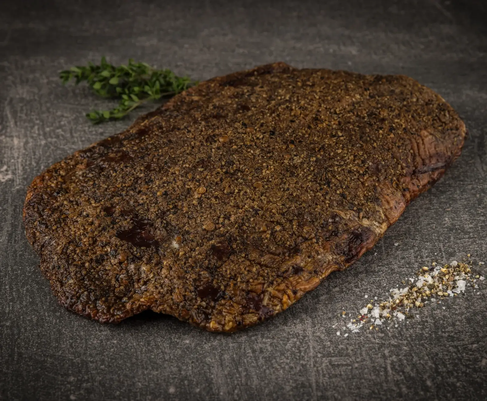 Ein großes, dunkelbraunes Stück Fleisch, reichlich mit grobem Pfeffer gewürzt, liegt auf einer dunklen Oberfläche. Ein Thymianzweig und eine Prise Salz und Pfeffer sind daneben.