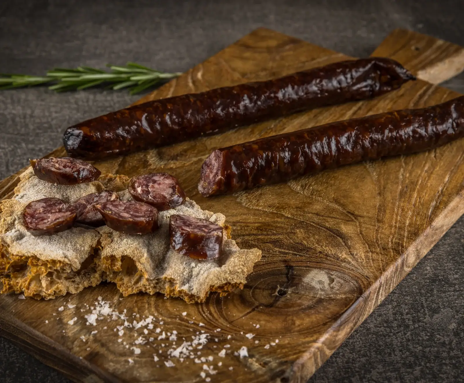 Zwei dunkle, glänzende Würste auf einem Holzschneidebrett. Eine Wurst ist angeschnitten, daneben liegen Scheiben auf rustikalem Brot mit Salzkristallen. Ein Rosmarinzweig ist im Hintergrund.